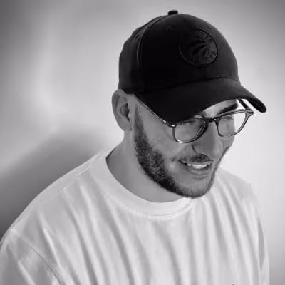 Man with a beard wearing glasses, a black baseball cap, and a white shirt smiling while looking down against a plain background.